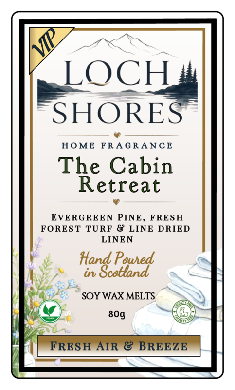 Front-facing product label for Loch Shores Home Fragrance 'The Cabin Retreat' soy wax melts, with a cream background, black and gold typography, mountain and lake artwork, floral illustrations, and a stack of folded white cloths at the bottom right.