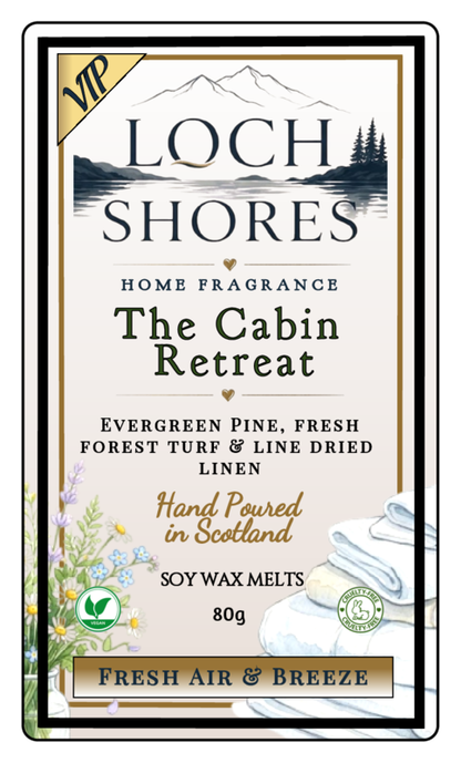 Front-facing product label for Loch Shores Home Fragrance 'The Cabin Retreat' soy wax melts, with a cream background, black and gold typography, mountain and lake artwork, floral illustrations, and a stack of folded white cloths at the bottom right.