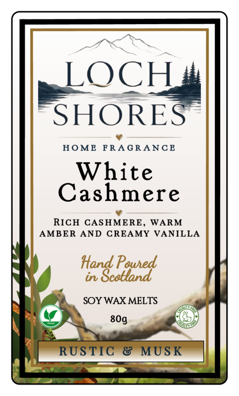Front-facing product label for Loch Shores White Cashmere home fragrance soy wax melts, with scenic mountain and lake artwork, text describing the scent as rich cashmere, warm amber, and creamy vanilla, and a Rustic & Musk note at the bottom.